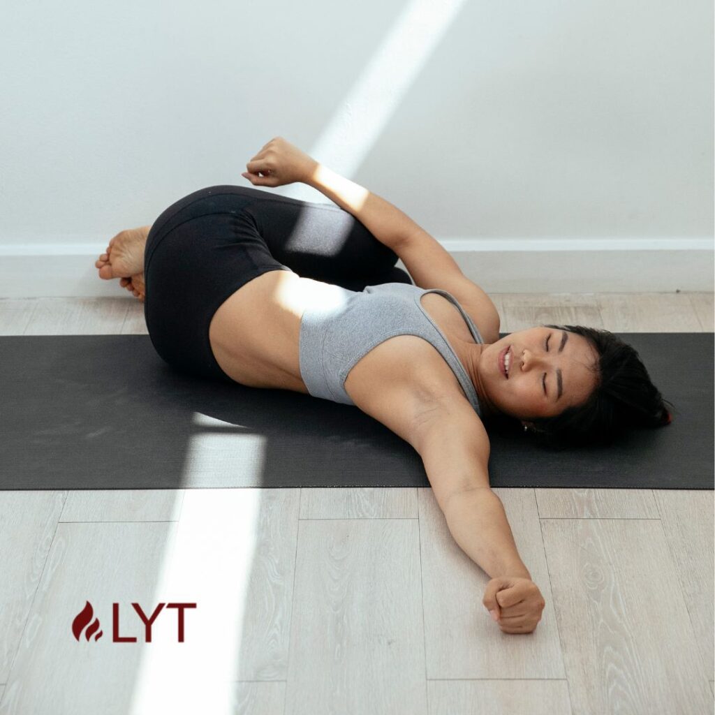 A woman performing the reclining spinal twist pose in yoga at her home over a black yoga mat