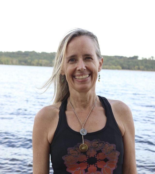 Smiling blonde woman wearing a mandala-print tank top, standing by a lake.