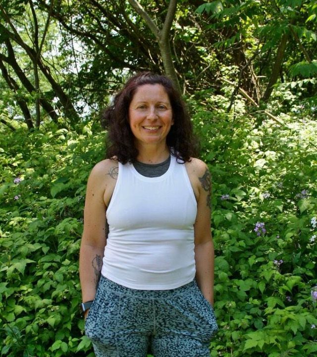 Smiling woman with curly hair standing outdoors surrounded by lush green foliage.